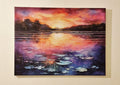 A rectangular canvas painting of a sunset over a lake with lily pads, shown hanging on a beige wall. The artwork features a vibrant orange and pink sky reflecting onto the water, with dark silhouetted trees and purple-blue water tones.