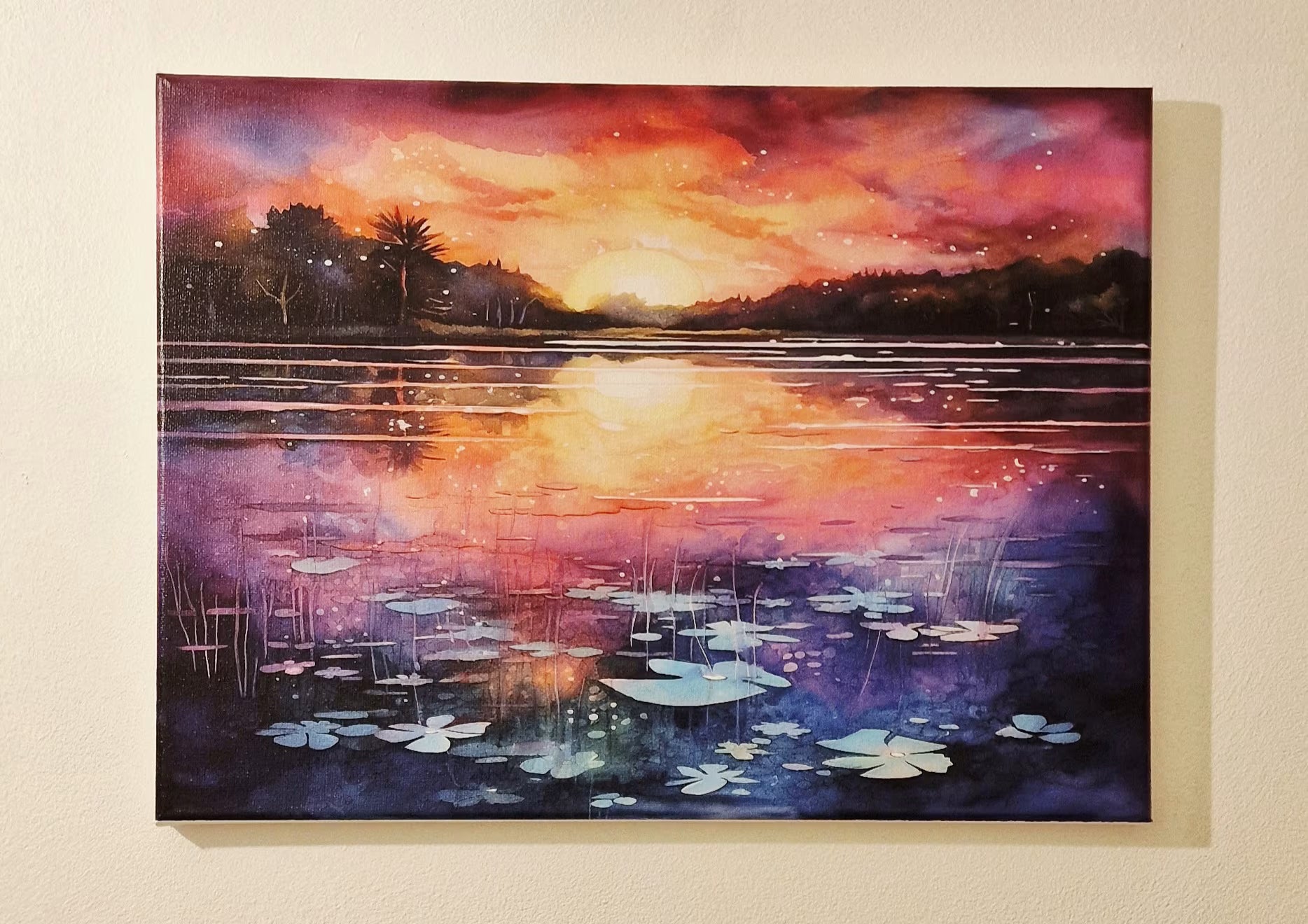 A rectangular canvas painting of a sunset over a lake with lily pads, shown hanging on a beige wall. The artwork features a vibrant orange and pink sky reflecting onto the water, with dark silhouetted trees and purple-blue water tones.