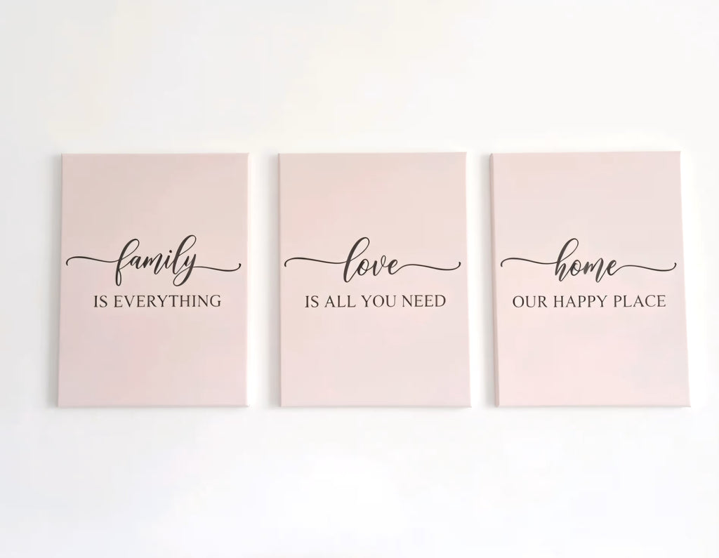 Three vertical canvas wall art prints in a soft beige color, arranged side by side on a white wall. Each print features elegant black script and serif text: 'family IS EVERYTHING', 'love IS ALL YOU NEED', and 'home OUR HAPPY PLACE'.