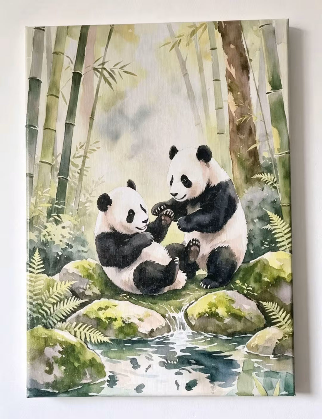 A watercolor painting of two pandas sitting on mossy rocks in a bamboo forest with a small stream in the foreground, shown as a square canvas print against a white wall.