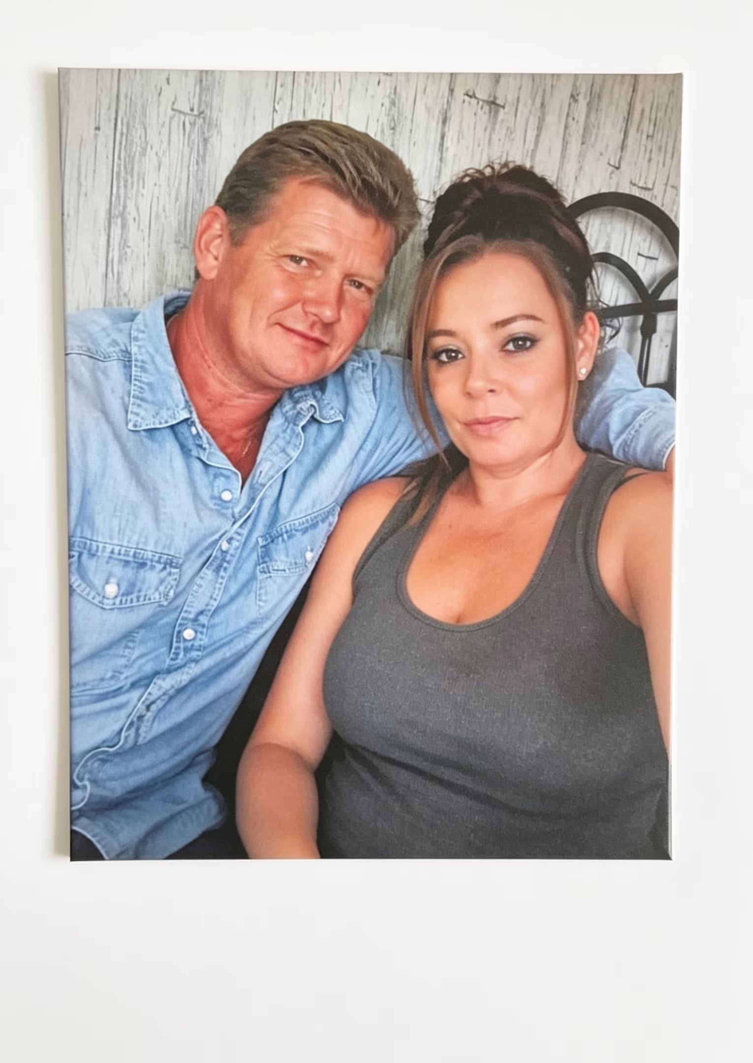 A square custom photo print of a smiling couple, displayed on a clean white wall. The man on the left wears a light blue denim shirt, and the woman on the right wears a dark gray tank top.