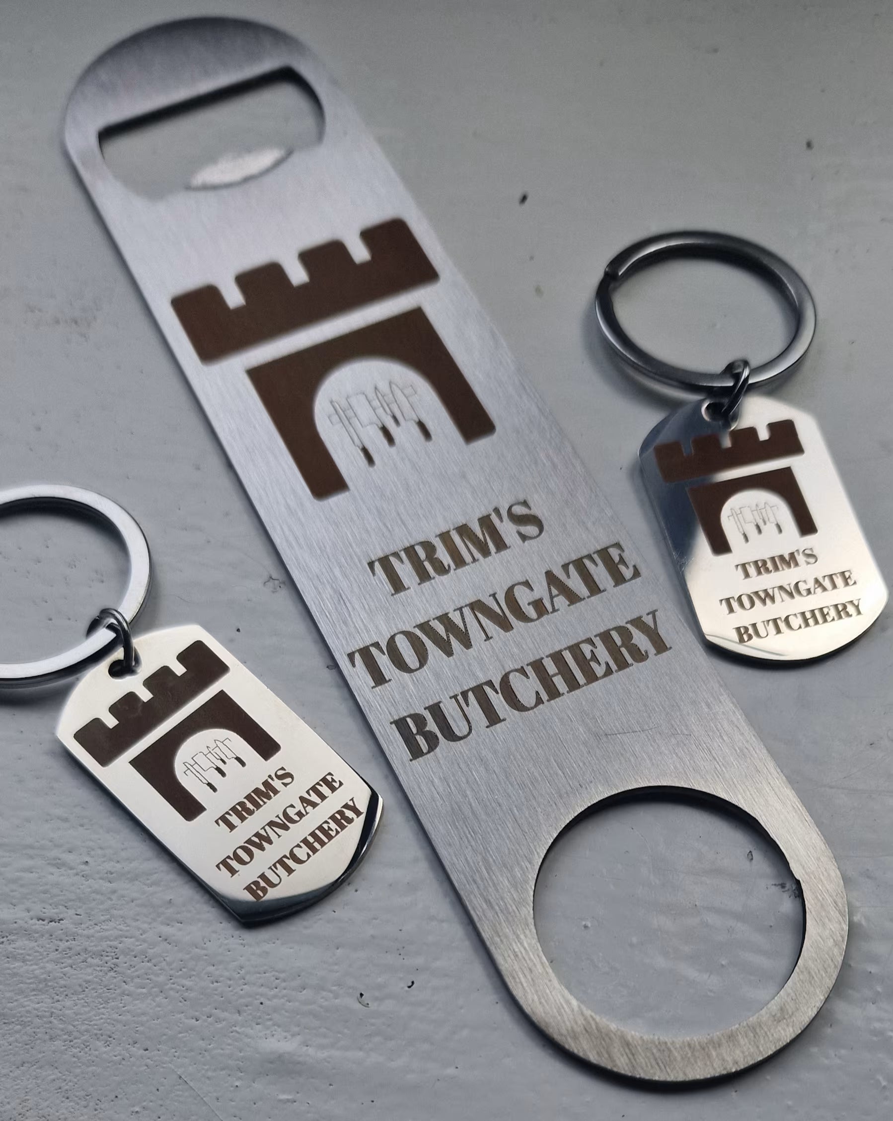 Three metal keychain-style bottle opener tools with engraved branding and a castle icon, arranged on a gray textured surface. The center item is a long rectangular opener, with two smaller oval keychain openers on either side.