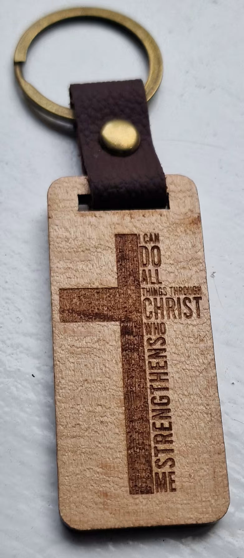 A wooden rectangular keychain with a brown cross engraving and the text 'I CAN DO ALL THINGS THROUGH CHRIST WHO STRENGTHENS ME', attached to a dark brown leather loop with a brass snap and gold-tone key ring, shown on a light textured background.